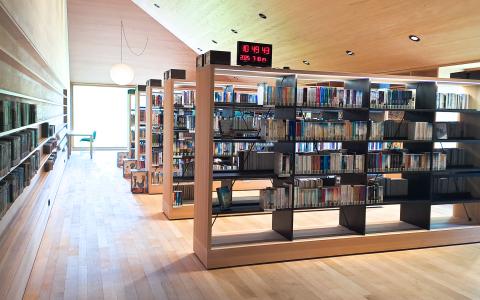 Bücherei innen - Gang mit Regalen an der Wand links und Regalen im Raum im 90 Grad Winkel zur Wand rechts vom Gang
